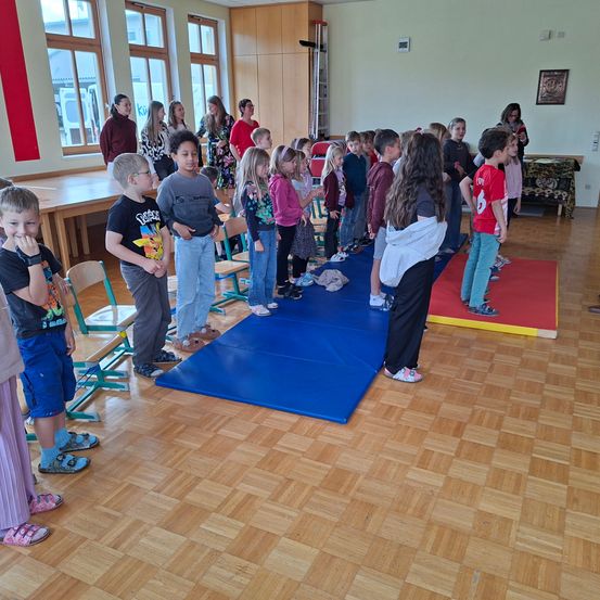Eine Gruppe von Kindern und Erwachsenen steht in einem Klassenzimmer. Einige stehen auf einem blauen Teppich, andere in der Nähe des Fensters oder in einer Ecke.