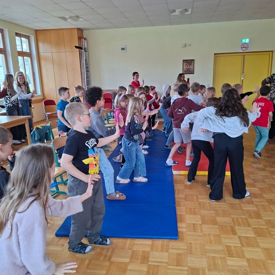 Eine Gruppe von Kindern tanzt auf einem blauen Teppich in einem Klassenzimmer, einige tragen Brillen und Turnschuhe. Einige Erwachsene schauen zu.