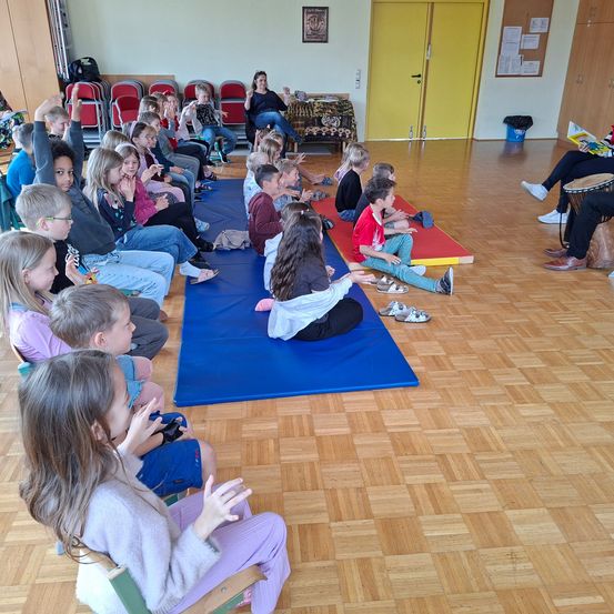 Eine Gruppe von Kindern sitzt auf einer blauen Matte in einem Raum und blickt auf eine Frau, die eine Sitzung leitet. Einige Kinder klatschen. Der Raum hat Holzböden und gelbe Türen.
