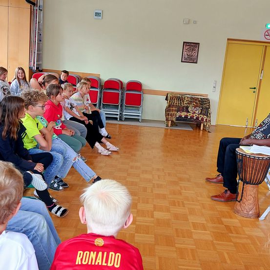 Eine Gruppe von Kindern und Erwachsenen sitzt auf Stühlen und hört einer Person zu, die eine Trommel spielt. Der Raum hat Holzböden, rote Stühle und eine gelbe Tür.