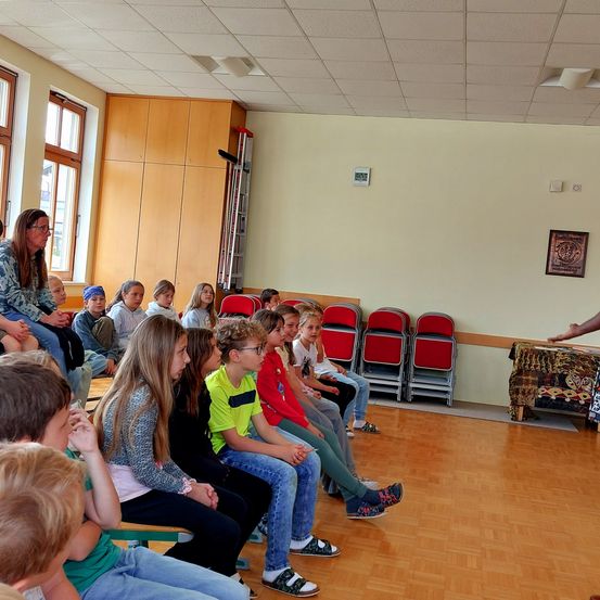 Ein Klassenzimmer mit Kindern, die sitzen, und einer Frau, die vorne steht, möglicherweise unterrichtet. Eine rote Leiter steht an der Wand, und rote Stühle sind in der Ecke aufgestellt. Ein Bilderrahmen hängt an der Wand.