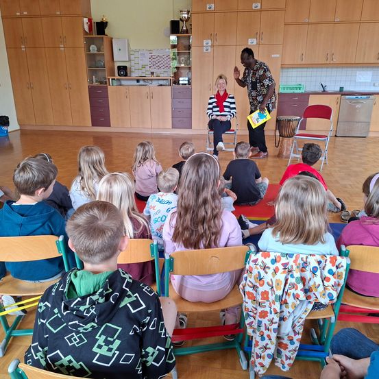 Ein Mann und eine Frau stehen vor einer Gruppe von Kindern in einem Klassenzimmer. Der Mann hält ein Buch und eine Trommel, während die Frau auf einem Stuhl sitzt. Die Kinder sitzen auf Stühlen und lauschen aufmerksam. Der Raum hat Holzböden und Schränke.