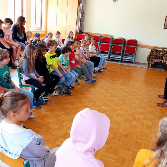 Kinder in einem Klassenzimmer sitzen in Reihen, nach vorne gerichtet, mit einer Lehrerin in der Nähe. Der Raum hat Holzböden, rote Stühle und einen Tisch in der Ecke.
