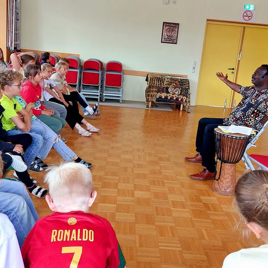 Eine Gruppe von Kindern sitzt in Stühlen um einen Mann, der eine Trommel spielt. Der Mann spricht zu ihnen. Die Kinder tragen verschiedenfarbige Shirts. Der Raum hat Holzböden und rote Stühle.