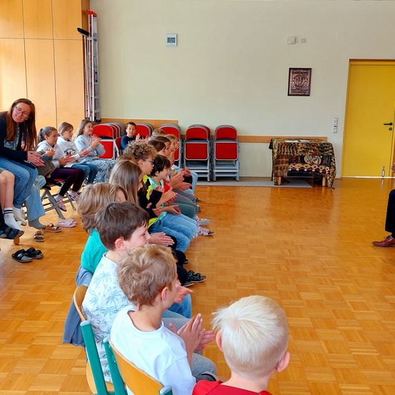 Eine Gruppe von Kindern und einer erwachsenen Frau sitzen auf Stühlen in einem Raum. Sie scheinen jemandem zuzuhören. Der Raum hat einen Holzboden, und eine gelbe Tür befindet sich rechts. Ein Rahmen ist an der Wand montiert. Ein Tisch steht in der Ecke des Raumes.