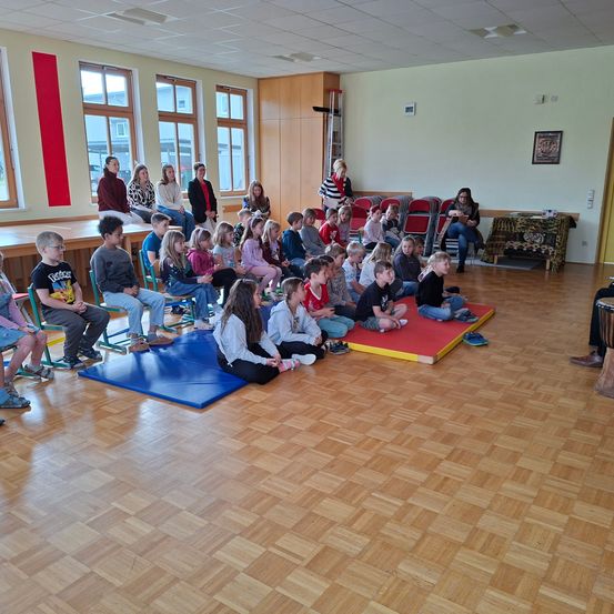 Eine Gruppe von Kindern sitzt im Kreis auf dem Boden, während Erwachsene in der Nähe stehen oder sitzen. Der Raum ist gut beleuchtet mit großen Fenstern und einem Bild an der Wand.