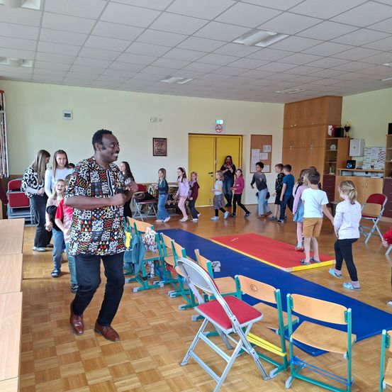 Ein Mann führt eine Gruppe von Kindern in einer Turnhalle an, wobei einige auf einem roten Teppich stehen, andere gehen und einige Stühle angeordnet sind. Ein Rauchverbotsschild ist sichtbar.