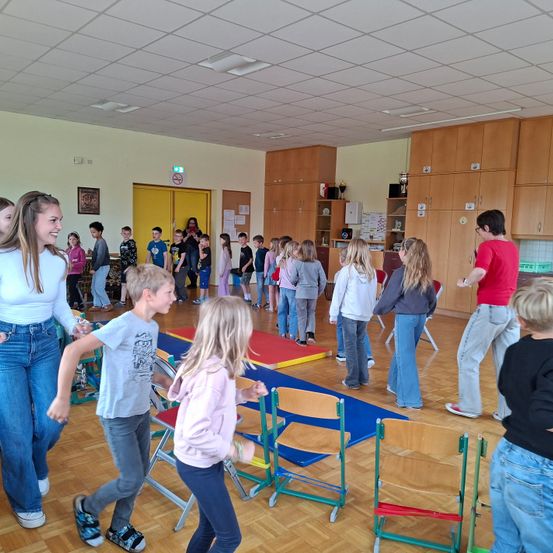Kinder spielen ein Spiel mit Stühlen in einem Klassenzimmer, einige stehen und einige sitzen. Eine Frau und ein Mann leiten die Aktivität.