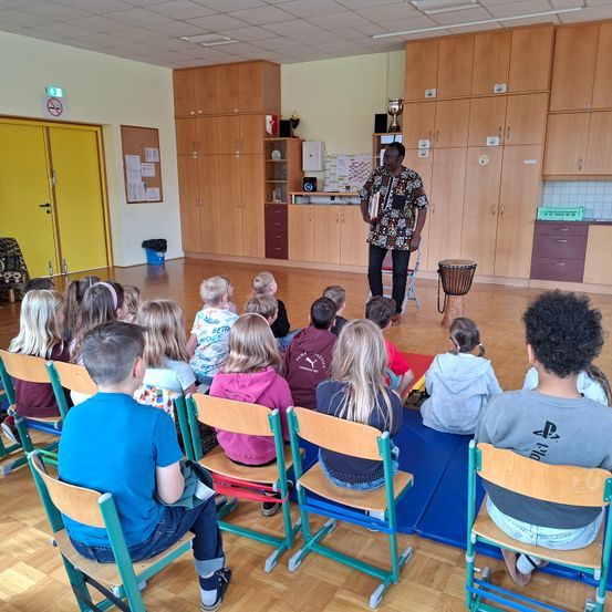 In einem Klassenzimmer hält ein Mann Bücher, während Kinder auf Stühlen vor ihm sitzen. Hinter ihm befinden sich Holzschränke und Regale. Ein Schlagzeug steht auf dem Boden in der Nähe der Schränke.