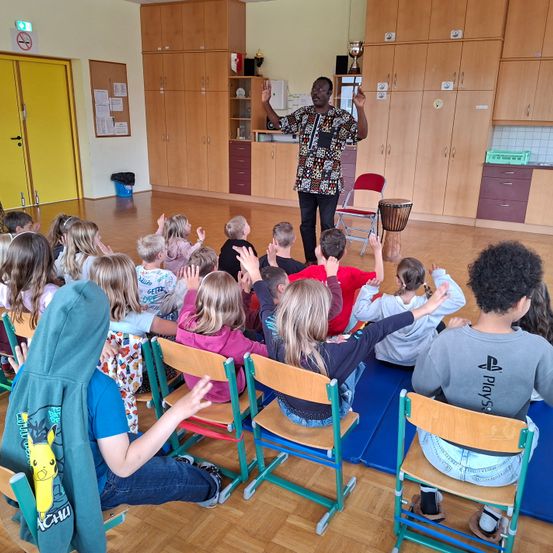 Ein Mann unterrichtet eine Klasse von Kindern. Sie sitzen in Reihen und hören aufmerksam zu. Der Raum hat Holzböden, Holzschränke und eine gelbe Tür.