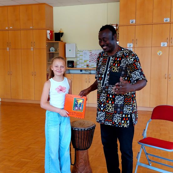 Ein lächelnder erwachsener Mann und ein junges Mädchen stehen neben einem Schlagzeug. Das Mädchen hält ein orangefarbenes Buch. Der Mann lächelt das Mädchen an.