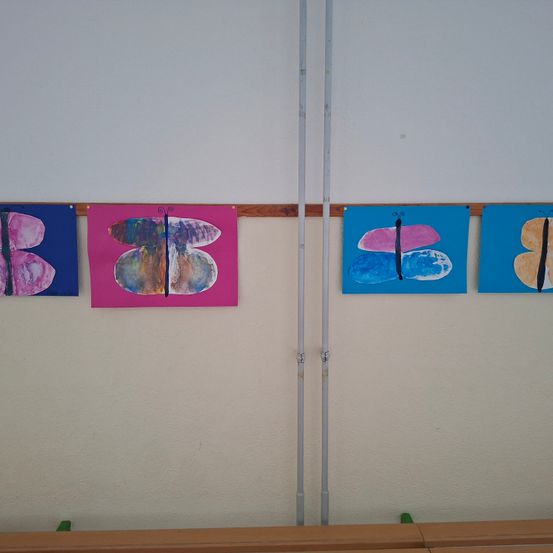 Vier Schmetterlingsgemälde hängen an einer Wand, jeweils in verschiedenen Farben und Stilen. Die Wand ist weiß und zwischen den Gemälden befindet sich ein Metallpfosten.