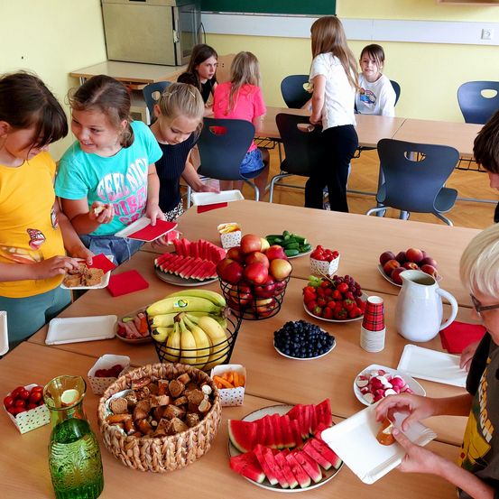 Eine Gruppe von Kindern versammelt sich um einen Tisch voller verschiedener Früchte, Gemüse und Snacks in einem Klassenzimmer.