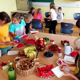 Eine Gruppe von Kindern versammelt sich um einen Tisch voller verschiedener Früchte, Gemüse und Snacks in einem Klassenzimmer.