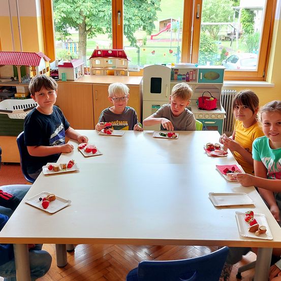 Fünf Kinder, drei Jungen und zwei Mädchen, sitzen um einen Tisch herum und essen von Tellern. Sie sind in einem Spielzimmer mit Spielzeug und einem Spielplatz, der durch die Fenster zu sehen ist.