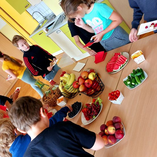 Kinder essen Snacks um einen Tisch mit Obst, Gemüse und anderen Lebensmitteln. Ein paar Teller mit Obst stehen auf dem Tisch.