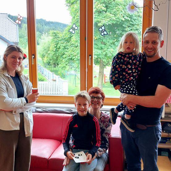 Eine Familie steht lächelnd in einem Wohnzimmer, mit einem kleinen Mädchen auf der Schulter eines Mannes. Sie haben einen Becher, ein rotes Sofa und ein Fenster mit Schmetterlingen.