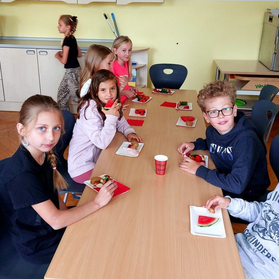 Eine Gruppe von Kindern sitzt am Tisch und isst. Ein Mädchen isst Wassermelone. Der Junge neben ihr trägt eine Brille.
