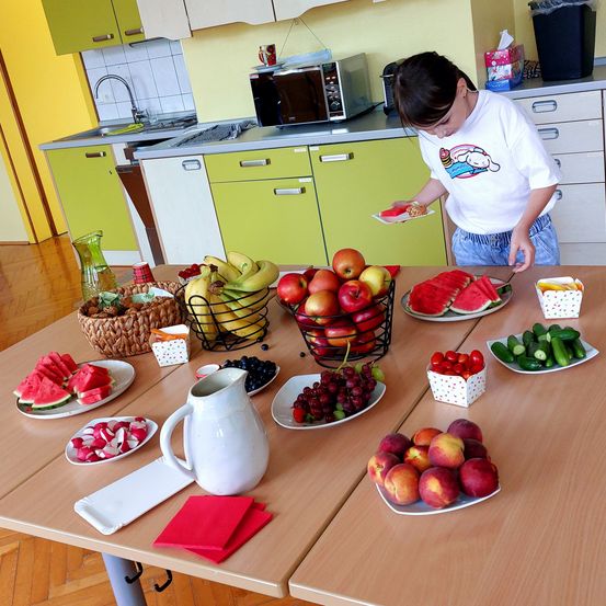Ein Kind in einer Küche serviert Obst und Gemüse auf einem Tisch, darunter Äpfel, Wassermelonen, Trauben und Radieschen.
