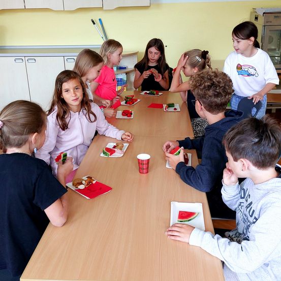 Eine Gruppe von Kindern sitzt um einen Tisch und isst Wassermelone. Einige haben die Hände erhoben. Der Tisch hat Teller mit Wassermelone und einen roten Becher.