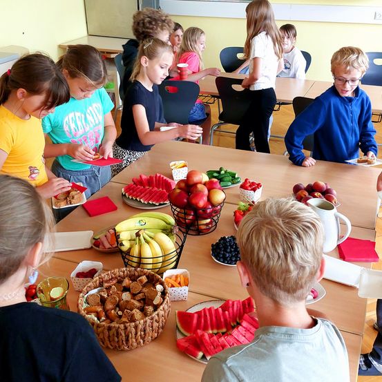 Eine Gruppe von Kindern versammelt sich um einen Tisch, der mit verschiedenen Früchten und Snacks gefüllt ist. Einige sitzen, andere stehen, und ein Junge mit Brille beugt sich über einen Teller mit Essen.