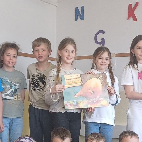 Sechs Kinder posieren für ein Foto, eines davon hält ein Buch mit dem Titel Das Osterkücken. Sie tragen alle Freizeitkleidung. Dahinter hat eine Wand große Buchstaben, die NKG ergeben.