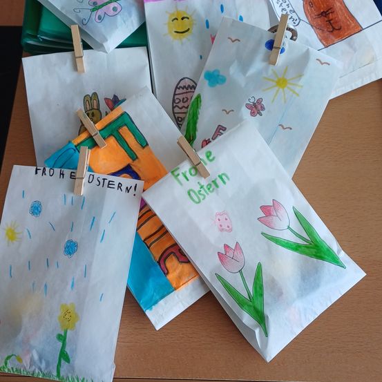 Mehrere handgemachte Ostertüten sind auf einem Tisch angeordnet. Jede Tüte ist mit Zeichnungen von Blumen, Schmetterlingen und Häusern verziert. Sie sind mit Wäscheklammern befestigt.