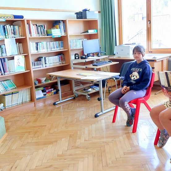 Ein Mädchen sitzt auf einem roten Stuhl in einem Klassenzimmer, umgeben von hölzernen Bücherregalen und Schreibtischen. Ein Computer-Setup befindet sich auf einem Schreibtisch, und ein Drucker ist in der Nähe.