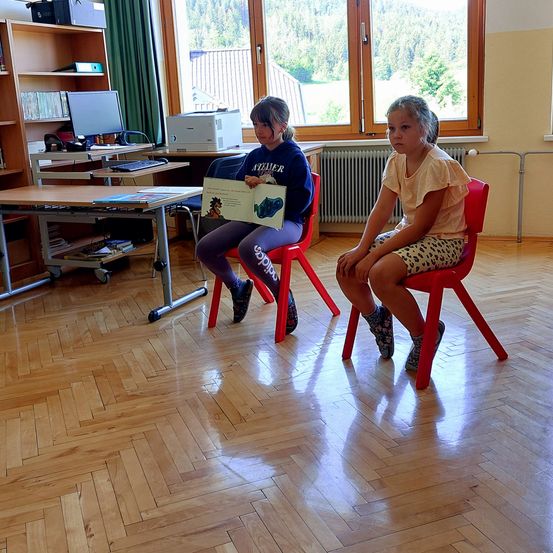 Zwei Mädchen sitzen auf roten Stühlen. Eine liest aus einem Buch. Hinter ihnen steht ein Schreibtisch mit Computer, Drucker und Büchern. Ein Regal steht hinter dem Computer. Fenster mit Vorhängen bieten einen Blick auf einen Berg.