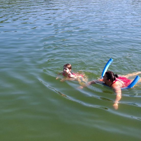 Zwei Personen, eine Frau und ein Kind, schwimmen rücklings im ruhigen Wasser und nutzen Schwimmhilfen zur Unterstützung. Das Wasser ist klar mit sanften Wellen.