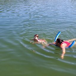 Zwei Personen, eine Frau und ein Kind, schwimmen rücklings im ruhigen Wasser und nutzen Schwimmhilfen zur Unterstützung. Das Wasser ist klar mit sanften Wellen.