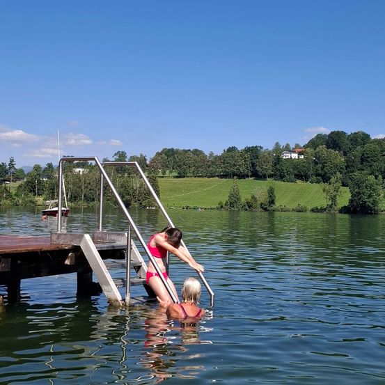 Zwei Frauen sind in einem See, eine klettert eine Leiter hoch, während die andere vollständig untergetaucht ist. Ein Boot ist links angedockt.
