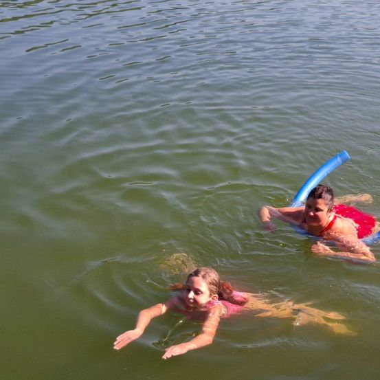 Zwei Mädchen treiben auf dem Rücken im Wasser und benutzen blaue aufblasbare Schwimmreifen. Das Mädchen vorne trägt einen pinkfarbenen Badeanzug und lächelt in die Kamera.