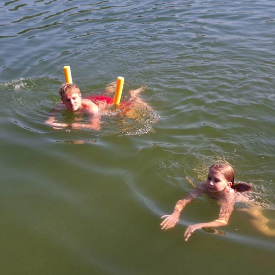 Zwei Kinder schwimmen in einem See und benutzen Schwimmnudeln als Schwimmhilfen. Ein Junge trägt einen roten Badeanzug, das Mädchen hat das Haar zurückgebunden.