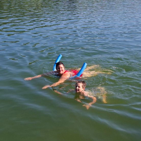 Zwei Menschen schwimmen auf blauen Schwimmhilfen im See und genießen das Wasser. Die Frau liegt auf dem Rücken, während das Kind in der Nähe schwimmt.