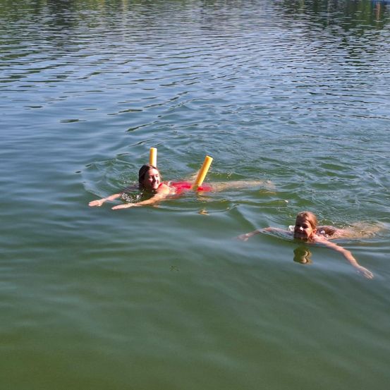 Zwei Mädchen schwimmen mit Schwimmhilfen in einem See. Sie lächeln und genießen ihre Zeit im Wasser.