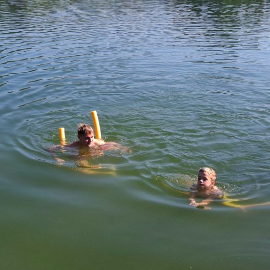 Zwei Jungen schwimmen in einem See mit gelben Schwimmhilfen. Ein Junge schaut zur Kamera, während der andere nach unten schaut.