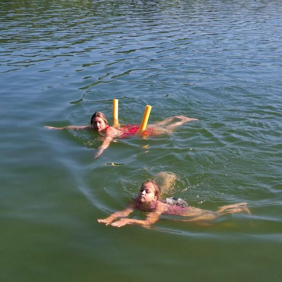 Zwei Kinder schwimmen in einem See, eines mit einem gelben Schwimmer. Das Wasser ist ruhig und der Himmel klar.