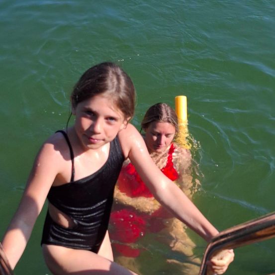 Zwei Mädchen in Badeanzügen sind in einem grünen Gewässer. Das Mädchen in Schwarz hält eine Handstütze, während das Mädchen in Rot mit einem Schnorchel schwimmt.