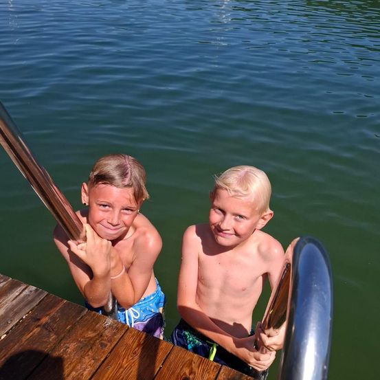 Zwei Jungen stehen auf einem Steg, halten sich an den Geländern fest, lächeln und schauen in die Kamera. Ein Junge hat blonde Haare, der andere braune Haare. Das Wasser ist ruhig.