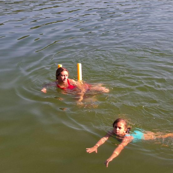 Zwei Mädchen schwimmen in einem See mit gelben Schwimmhilfen. Ein Mädchen trägt einen roten Bikini und das andere ein blaues Badekleid. Sie genießen das Wasser.
