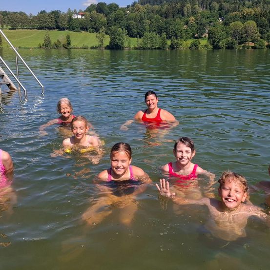 Eine Gruppe von Menschen schwimmt in einem See. Sie alle lächeln. Hinter ihnen führt eine Treppe zum Wasserrand. Das Gebiet ist von üppiger Grünfläche umgeben.