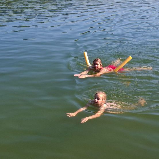 Zwei junge Mädchen treiben im ruhigen Wasser und nutzen gelbe Schwimmhilfen. Ein Mädchen hat rosa Schwimmhilfen, das andere gelbe.