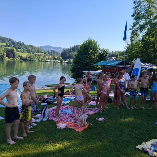 Eine Gruppe von Kindern und Jugendlichen versammelt sich auf einer Wiese in der Nähe eines Sees, einige in Badekleidung, bereit für eine Aktivität.