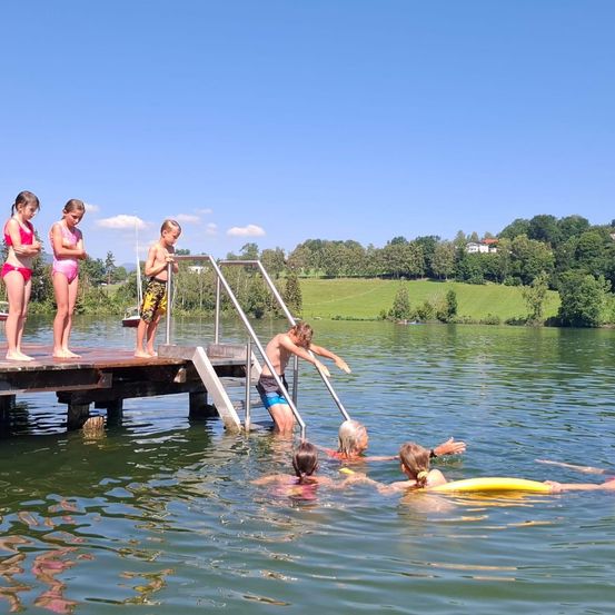 Mehrere Kinder und Erwachsene schwimmen und spielen in einem See in der Nähe eines hölzernen Stegs mit einer Metallhandlauf.
