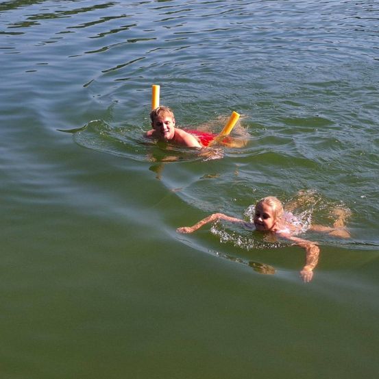Zwei Kinder schwimmen mit gelben Schnorcheln im Wasser. Ein Junge trägt rote Badeshorts. Sie schauen zur Kamera.
