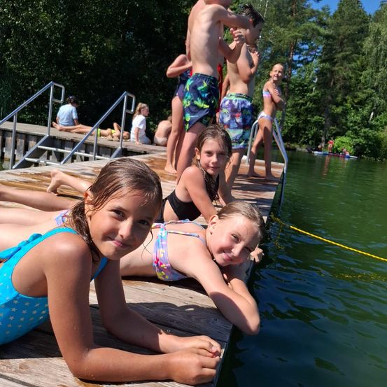 Eine Gruppe von Kindern und Jugendlichen auf einem hölzernen Steg an einem See. Einige liegen auf dem Steg, während andere am Rand stehen oder sitzen. Bäume und ein klarer Himmel sind im Hintergrund zu sehen.