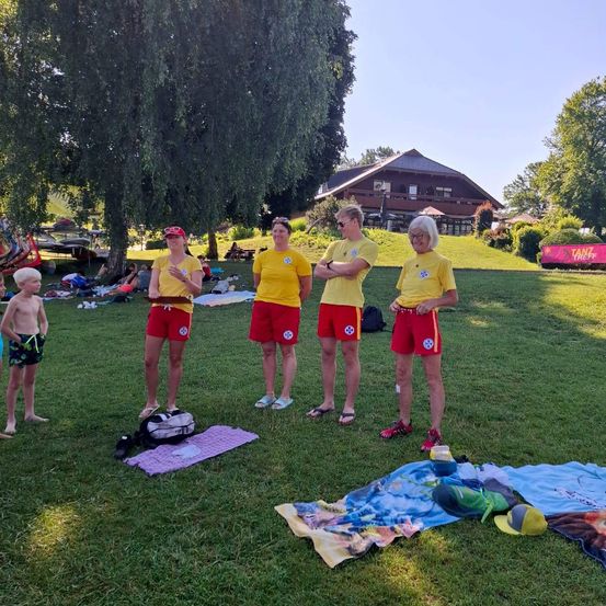 Eine Gruppe von Rettungsschwimmern in gelben Uniformen und roten Shorts steht auf einem Rasen, ein Kind ist in der Nähe. Ein Baum, ein Haus und ein Banner sind im Hintergrund.