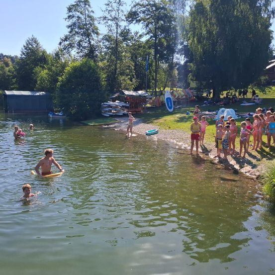 Eine Gruppe von Kindern und Erwachsenen genießt einen Tag am See. Einige schwimmen, andere stehen am Ufer. Es gibt auch Boote und Zelte in der Umgebung.