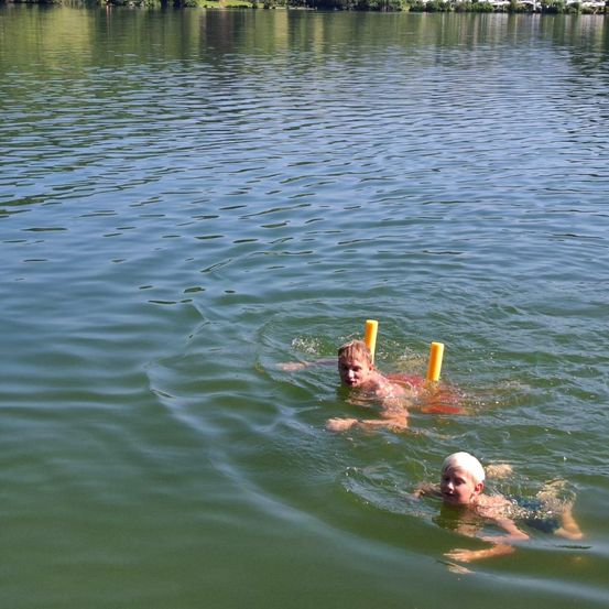 Zwei Jungen schwimmen in einem See, einer mit einem gelben Schwimmhilfe. Das Wasser ist ruhig und grün. Bäume und Gebäude sind im Hintergrund sichtbar.